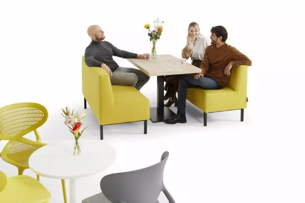 Drie lachende mensen zitten samen op een gele kantinebank aan tafel.